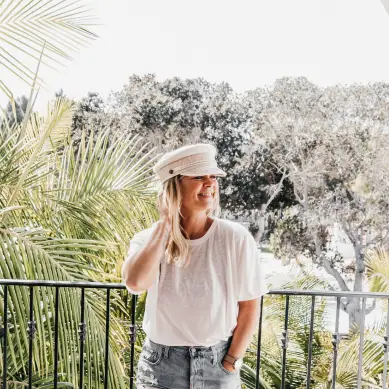 A person stands on a balcony surrounded by palm trees, wearing a light-colored t-shirt and a matching cap, with one hand touching their hair. The background features more greenery and a bright sky.