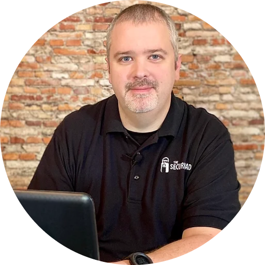 Man with short hair and a beard wearing a black polo shirt sits at a desk with a laptop. The shirt has The Securidad logo. Brick wall in the background.