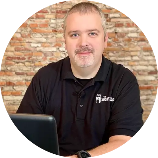 Man with short hair and a beard wearing a black polo shirt sits at a desk with a laptop. The shirt has The Securidad logo. Brick wall in the background.