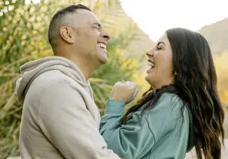 A man and a woman stand outdoors, laughing together. The man wears a beige hoodie, and the woman wears a green jacket. They are surrounded by greenery, with soft sunlight in the background, creating a warm and joyful atmosphere.