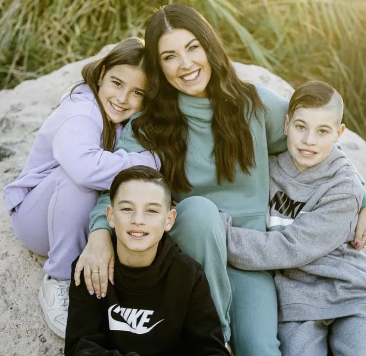 A woman sits on a rock with her arms around three children. They are all dressed in casual, sporty clothes, smiling at the camera. The background features green foliage and sunlight.