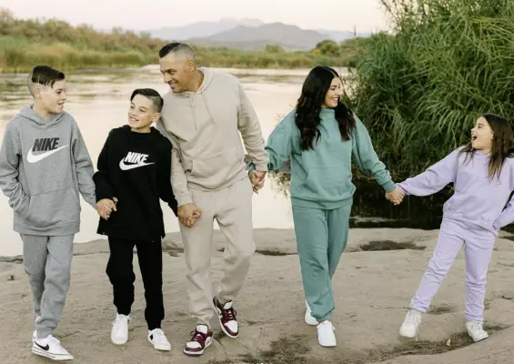 A family of five is walking hand in hand near a body of water, surrounded by greenery. They are wearing casual sportswear and smiling at each other. The background shows distant mountains under a pastel sky.