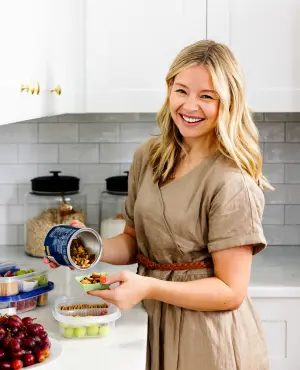 A woman in a beige dress stands in a kitchen, smiling while pouring almonds from a container into her hand. Nearby, there are containers of grapes and strawberries on a white countertop.