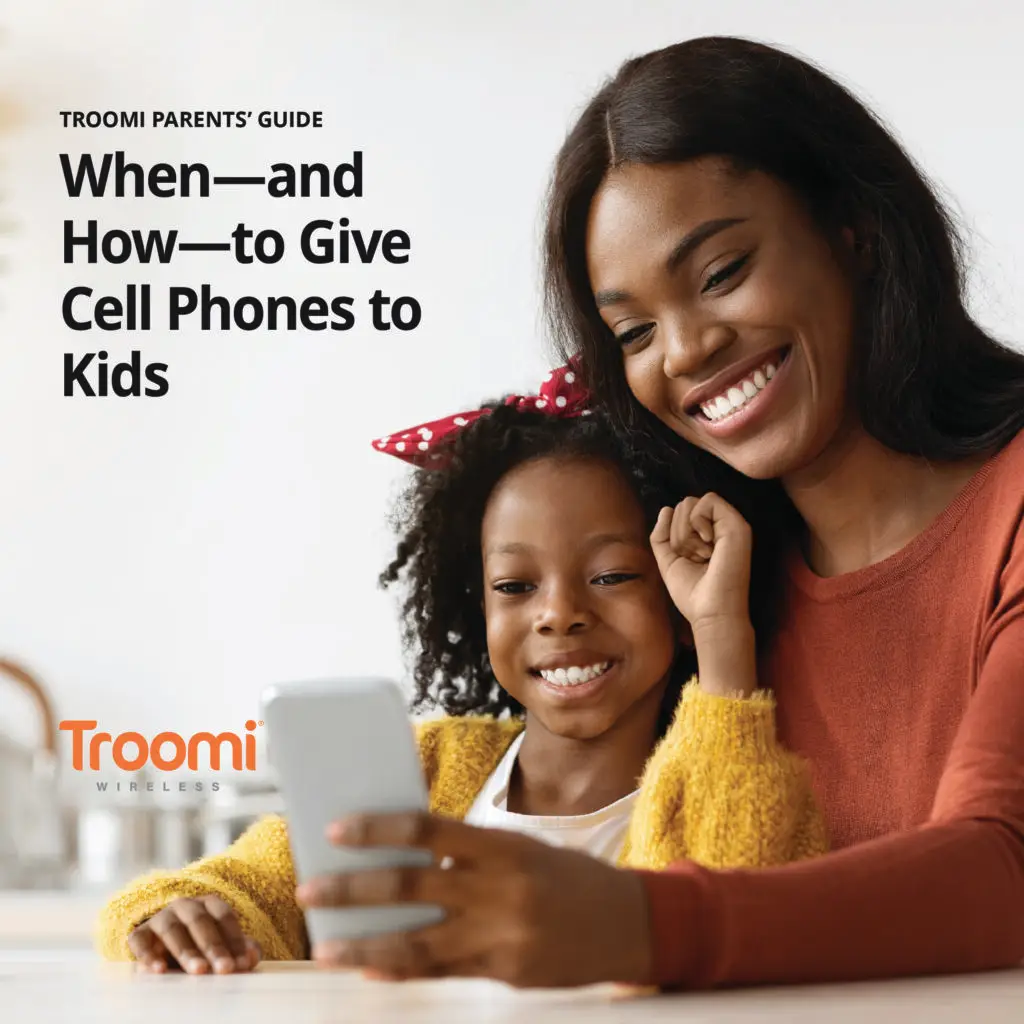 A smiling adult and child sit together, engrossed in a smartphone. Text on the image reads: Troomi Parents Guide: When—and How—to Give Cell Phones to Kids. Discover downloadable resources for parents. The Troomi Wireless logo is in the bottom left corner.