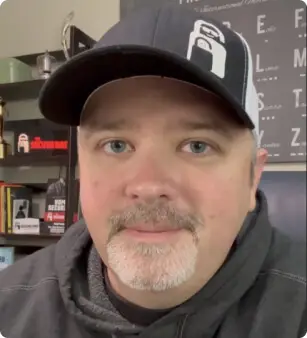 A man with a goatee wears a black baseball cap and a gray hoodie. He is indoors, with shelves holding books and trophies in the background. A chart or board is partially visible behind him.