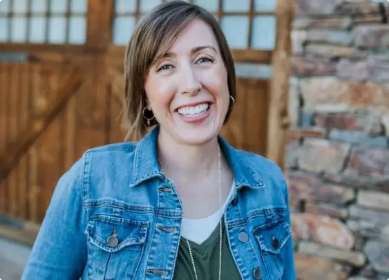 A person smiling outdoors, wearing a denim jacket over a green top. They are standing in front of a stone wall and a wooden door with glass panels.