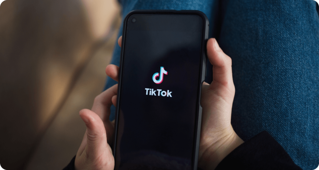 A person holds a smartphone displaying the TikTok logo on the screen, highlighting the app's popularity despite potential TikTok risks. In the background, a blurred figure casually sits in blue jeans and a dark top.