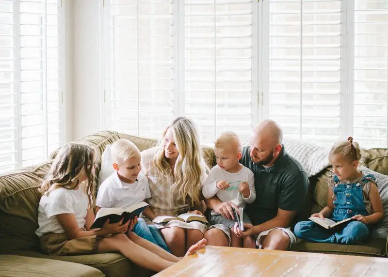 A family of six sits together on a cozy couch in a sunlit room, reading books. Three children hold books, while two adults and a toddler accompany them. They are surrounded by white shutters and a wooden table is in the foreground.