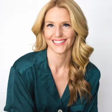 A person with long blonde hair wearing a teal shirt smiles widely with a white background.