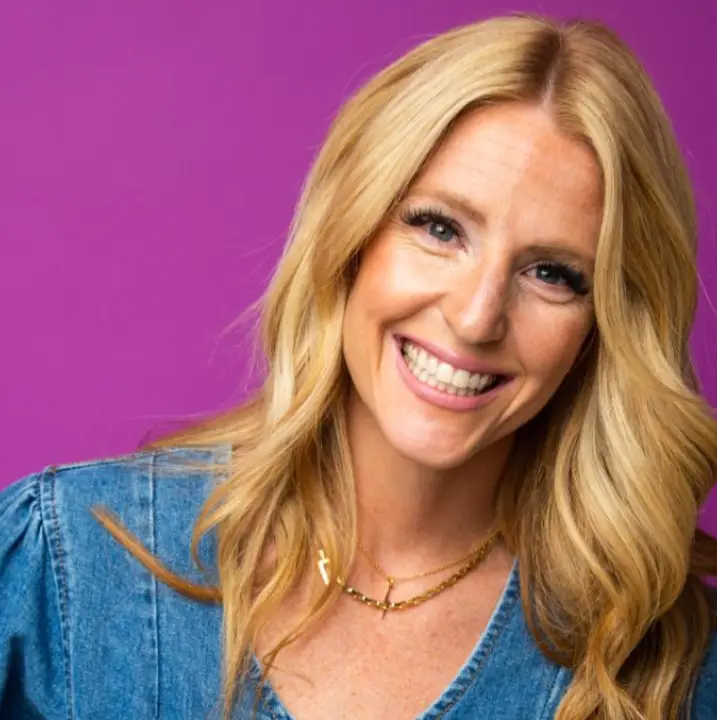 A smiling woman with long blonde hair is wearing a denim shirt and gold necklaces. She stands against a vibrant purple background.