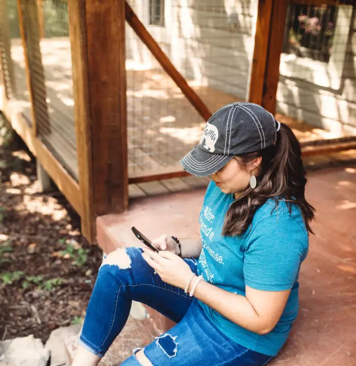 A person sitting on a porch step, wearing a black cap, blue T-shirt, and ripped jeans, is focused on a smartphone. The porch features wooden railings, and the setting is outdoors with sunlight filtering through.