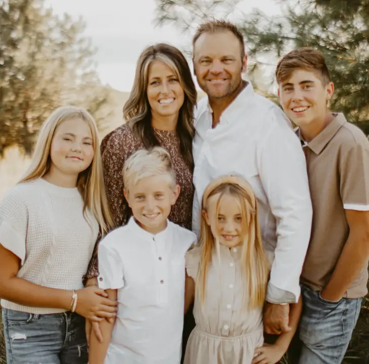 A family of six, including two adults and four children, poses outdoors. Everyone is smiling and dressed casually. They are surrounded by trees and grass, creating a warm and natural setting.