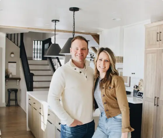 A couple stands smiling in a modern kitchen with white cabinets, wooden accents, and pendant lights. A staircase leads to the second floor in the background, and there are several decorative items on the counters.