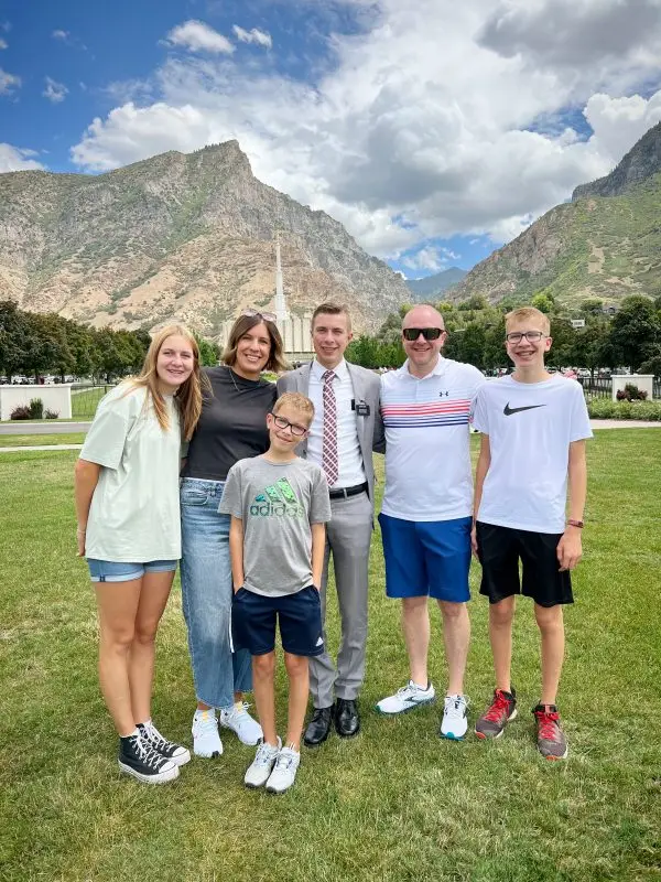 A group of six people smiling and standing on a grassy field with mountains and a cloudy sky in the background.