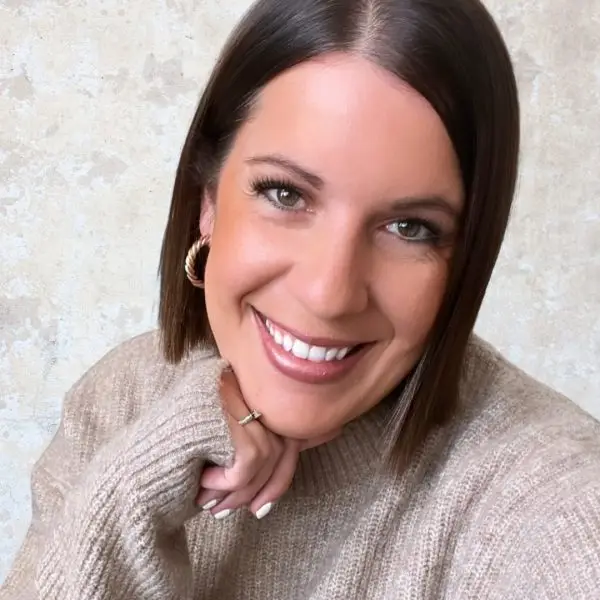 A woman with straight, shoulder-length brown hair smiles at the camera. She is wearing a beige sweater and hoop earrings, with her chin resting on her hand. The background is a light textured wall.