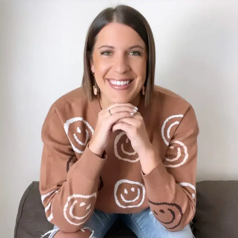 A person with short brown hair smiles warmly, resting their chin on their hands. They wear a brown sweater with white and brown smiley faces and light blue jeans. The background is plain white.