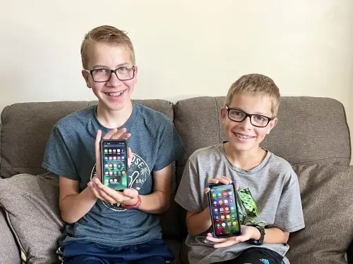 Two boys with glasses sit on a couch, smiling and holding smartphones with colorful app icons on the screens. They are wearing casual shirts and sitting against a light background.