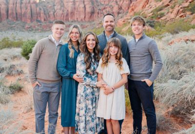 A family of six stands together in a desert landscape with red rocky cliffs in the background. They are dressed in casual, colorful outfits, smiling at the camera. The scene is sunny and serene, with sparse desert vegetation around them.