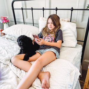 A girl is lounging on a bed with a black dog. She is focused on a smartphone, wearing a dark t-shirt and shorts. The bed has white bedding, and theres a pink flower in the background.