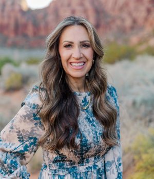 A woman with long wavy hair smiles at the camera, standing in a natural setting with red rock formations and greenery in the background. She is wearing a blue patterned dress.