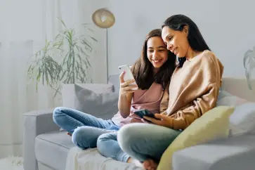 Two people sitting on a couch in a cozy living room. Both are smiling and looking at smartphones, appearing to share something interesting. A lamp and potted plant are in the background, adding a relaxed ambiance to the scene.