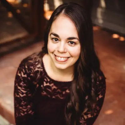A woman with long, dark hair and a lace top smiles while sitting on steps. The background is softly blurred, suggesting a cozy, outdoor setting.