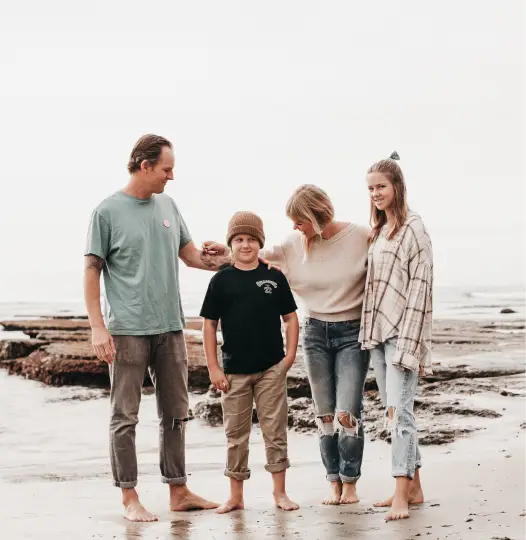 A family of four stands barefoot on a beach. The father, mother, young boy, and teenage girl are casually dressed. The mother affectionately rests her hand on the boys shoulder, and the others are smiling. The ocean and rocks are in the background.