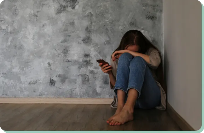 A person sits on the floor in the corner of a room, partially covering their face with one arm and holding a phone in the other hand. The background features a textured gray wall and wooden floor. The scene suggests a feeling of distress or contemplation.