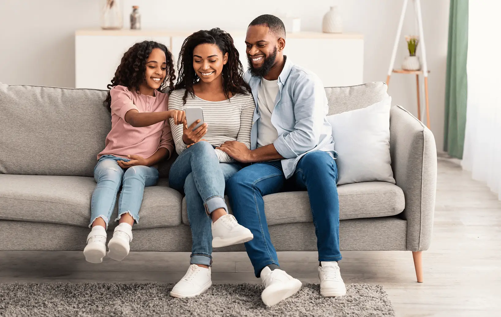 A family of three, a woman, a man, and a girl, sit on a sofa together, smiling and looking at a smartphone the girl is holding. The room is bright and modern, with light-colored decor and a cozy atmosphere.