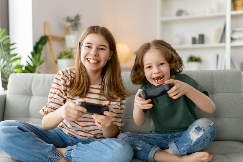 Two children sitting on a couch, smiling and holding game controllers. The older child wears a striped shirt and jeans, the younger child wears a green shirt and ripped jeans. A bookshelf and plants are visible in the background.