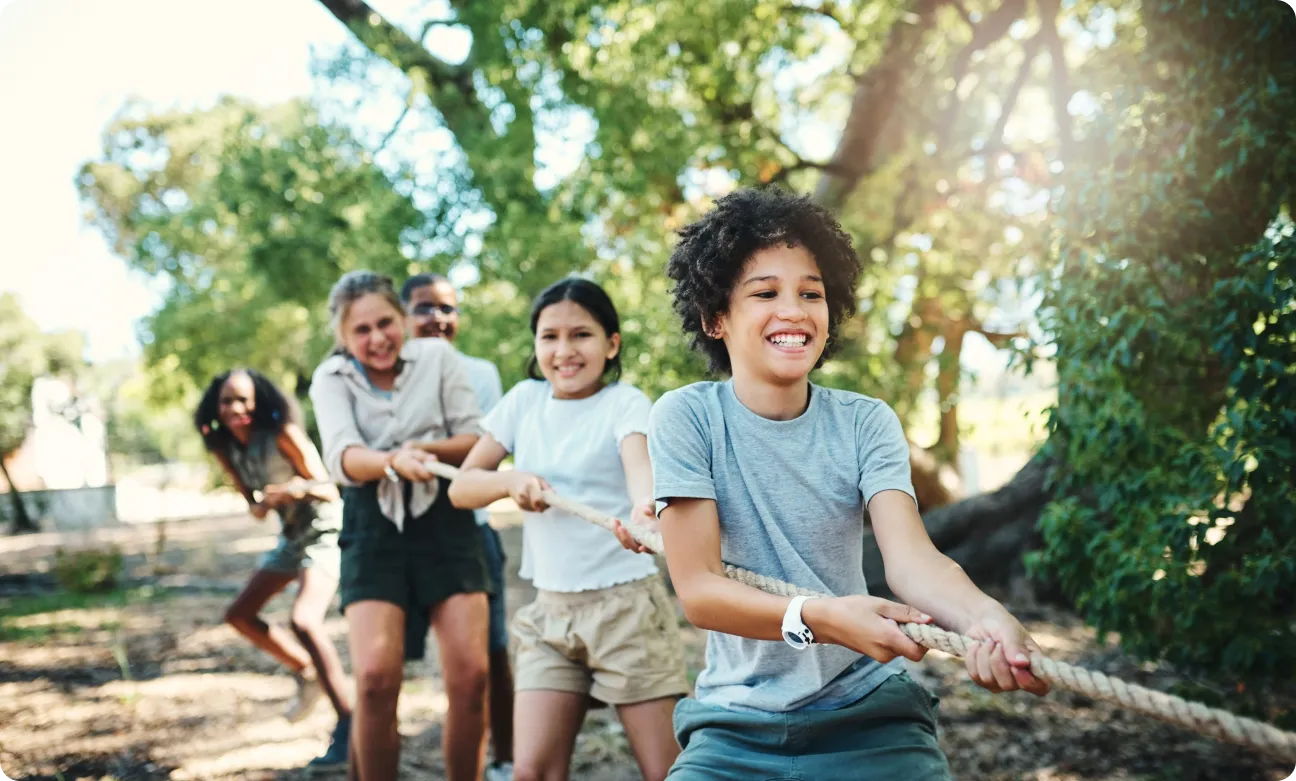 A group of smiling children playing tug-of-war outdoors. They are pulling on a rope with determination. Sunlight filters through the trees, creating a lively and playful atmosphere.