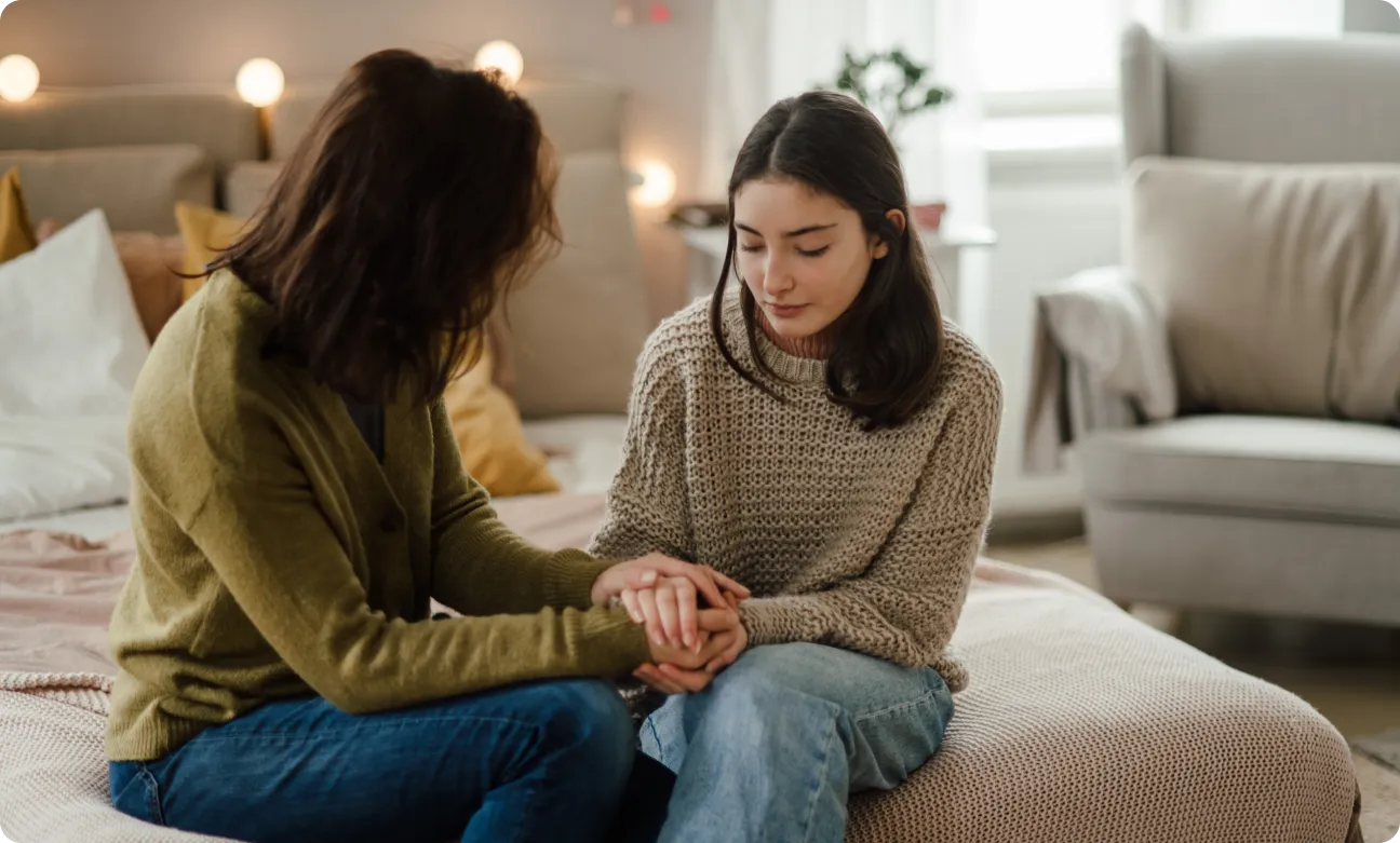 Two people sit on a bed in a cozy room, holding hands and looking at each other attentively. The room is softly lit, with decorative pillows and a comfortable chair in the background.