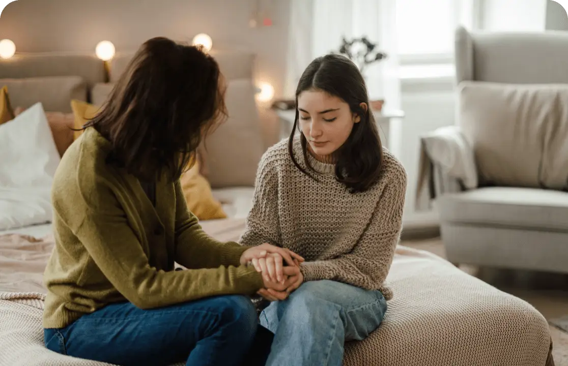 Two people are sitting on a bed facing each other, engaged in a supportive conversation, with one holding the others hands. The softly lit room, filled with cozy furnishings, feels inviting—a perfect setting to join the movement of deeper connections and understanding.