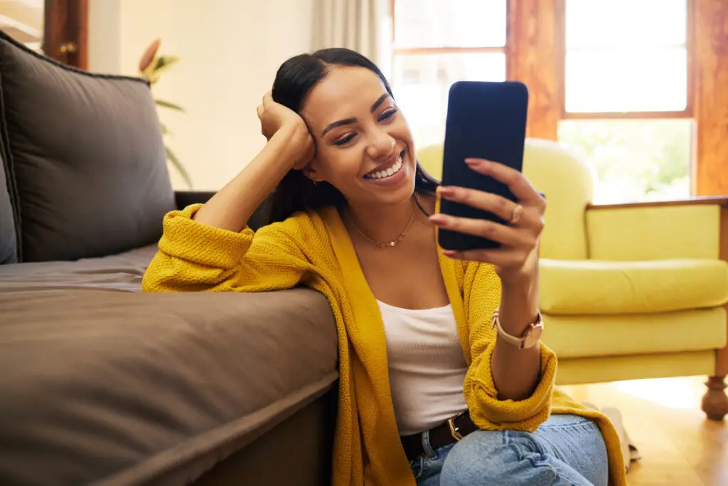 A woman in a yellow cardigan and white top sits on the floor, leaning against a couch. She is holding a smartphone and smiling. Sunlight filters through the window behind her, creating a warm, inviting atmosphere.