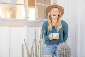 A woman with long blonde hair, wearing a hat and a teal long-sleeve shirt, smiles while holding a mug. She stands beside tall cacti in front of a white wall with a window, radiating warmth like an ambassador for cyberbullying prevention in this digital age.