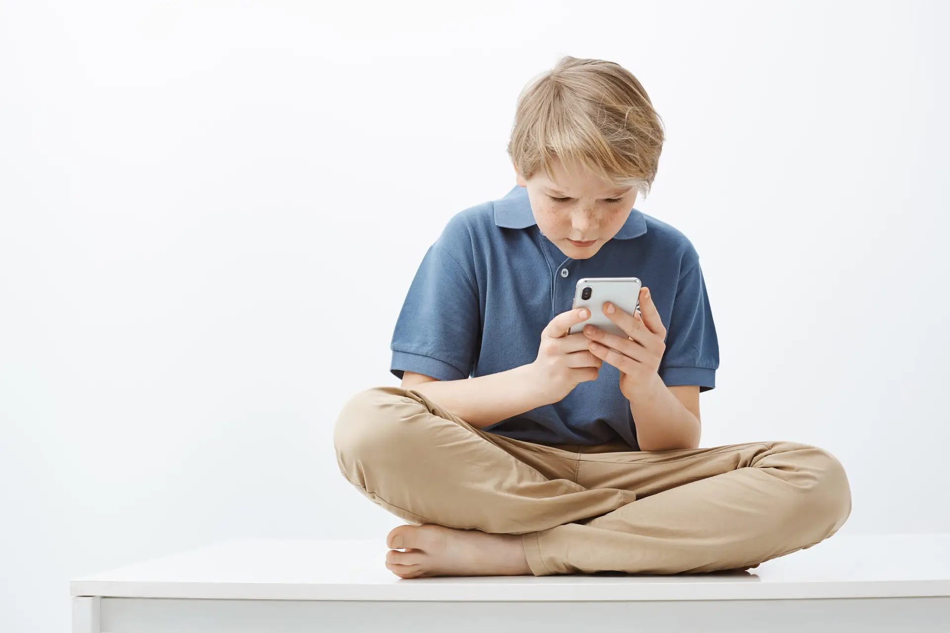 A young boy with blond hair sits cross-legged on a white surface, wearing a blue polo shirt and beige pants. He is intently looking at a smartphone he holds in both hands, perhaps participating in Safer Internet Day activities. The background is plain and light.