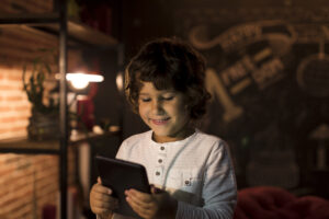 A smiling child with curly hair, wearing a light-colored long-sleeve shirt, uses a tablet in a dimly lit room. Shelves filled with books and a chalkboard adorned with drawings and text set the scene, highlighting the spirit of Safer Internet Day.