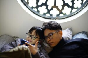 Two people lounging on a couch, looking at a phone together, possibly exploring tips for Safer Internet Day. One is smiling and wearing glasses, while the other leans against them. Behind them is a circular window with an intricate design.