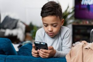 A young boy with short dark hair, wearing a light gray shirt, sits on a blue sofa, intently looking at a smartphone in his hands. In the spirit of Safer Internet Day, hes exploring digital safety tips. The background is blurred with indistinct decor and a hint of a TV screen.