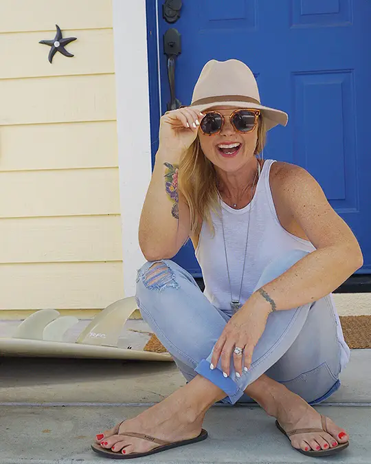 A woman wearing a beige hat, sunglasses, a white tank top, ripped jeans, and flip-flops sits casually on a porch. She is smiling and touching her hat brim, with a surfboard and blue door in the background.