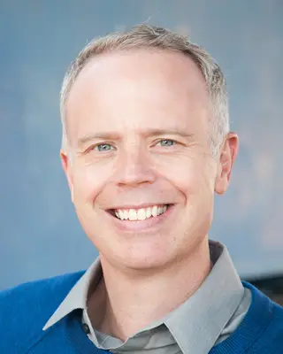 A smiling person with short, light-colored hair wearing a blue sweater over a gray collared shirt, embodying the spirit of cyberbullying prevention. The background is a soft, unfocused blue-gray.