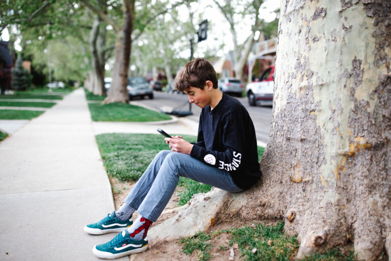 A young boy sits at the base of a large tree on a residential street, focused on his Troomi smartphone. Hes wearing a black long-sleeve shirt, gray pants, and teal sneakers. The background features parked cars and green grass swaying gently in the breeze.