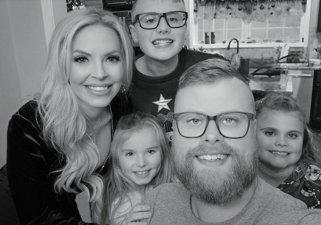 A smiling family of five poses for a photo indoors. From No Phone to Troomi, they enjoy time together, with an adult woman and man in front and three children—two girls and one boy—smiling around them. The background shows a kitchen and window.