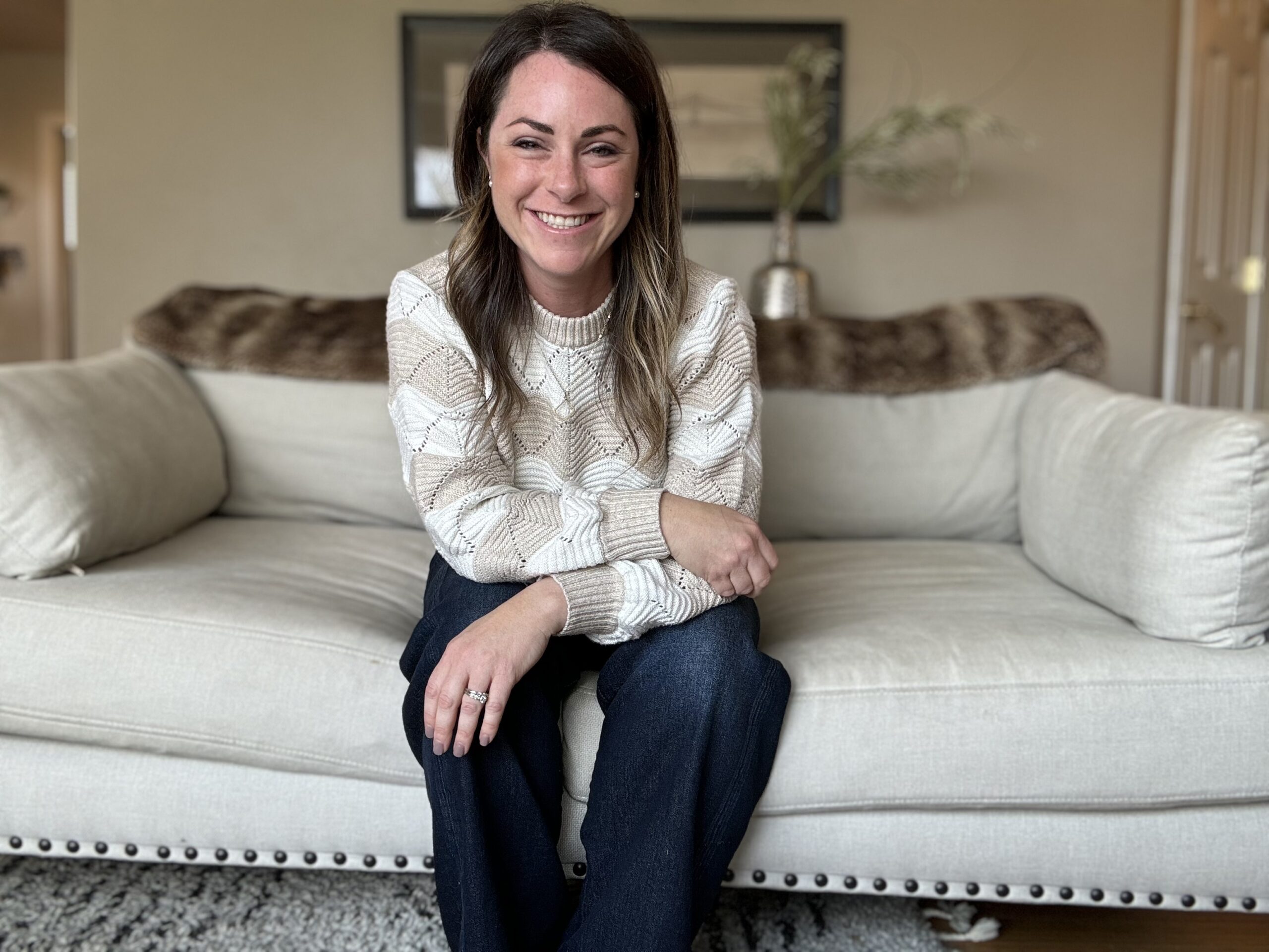 A woman with long brown hair sits smiling on a light-colored couch in a cozy living room. She is wearing a cream patterned sweater and dark pants, with her hands resting on her knee.