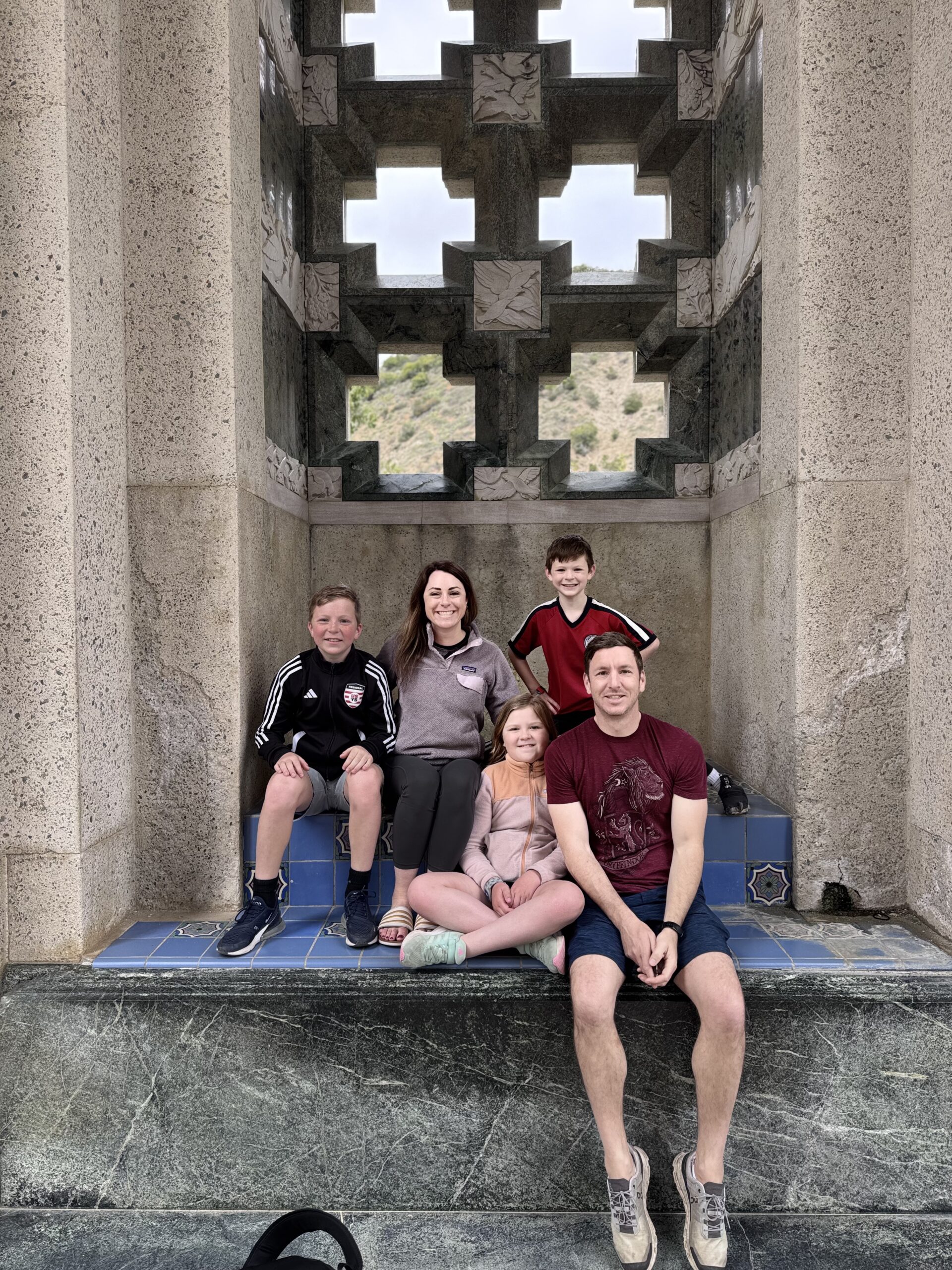 A family of five sits and kneels on blue-tiled steps inside a stone structure with geometric cutouts, smiling at the camera. The background shows patterned walls and a view through the windows.