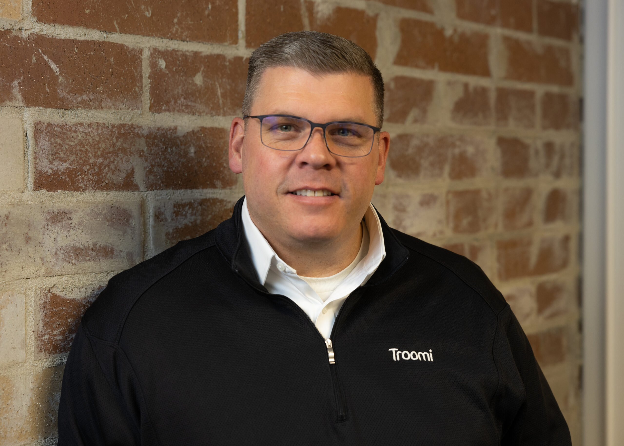 A man wearing glasses and a black jacket with the Troomi logo stands smiling against a brick wall.