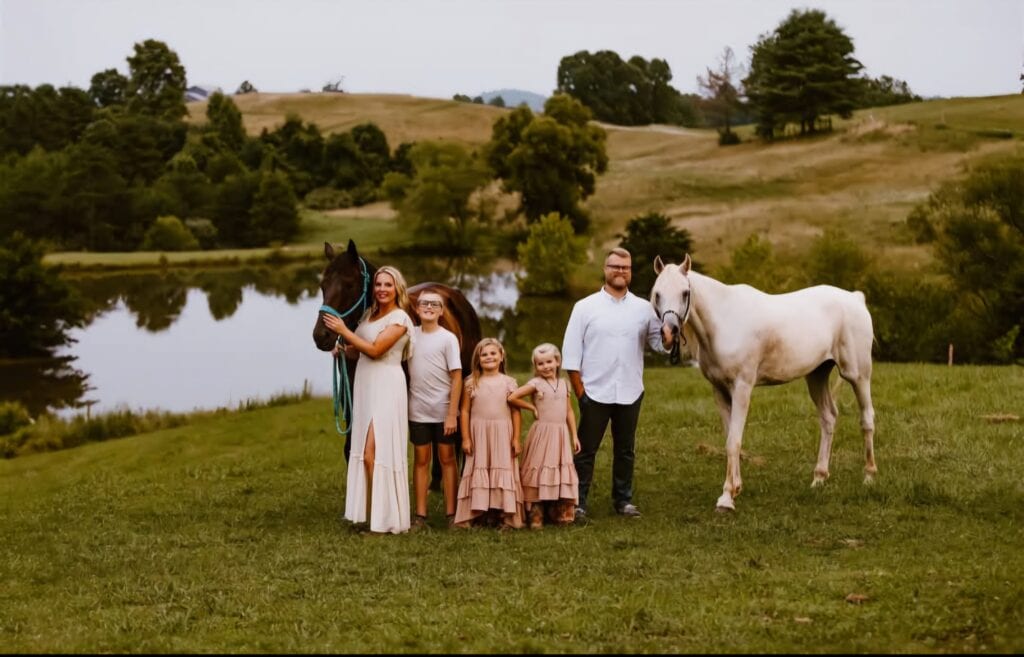 A family of five poses outdoors near a pond and rolling hills, standing between two horses. Smiling together on a grassy field under an overcast sky, they capture a From No Phone to Troomi moment in timeless, neutral tones.