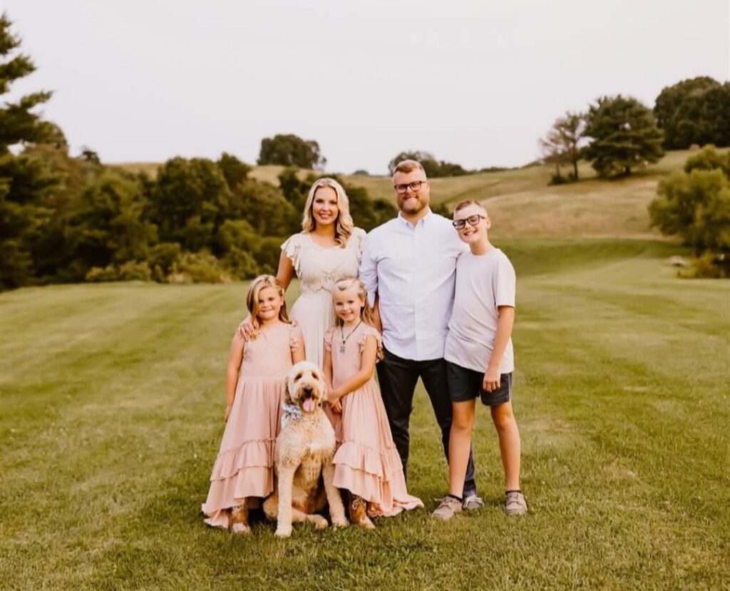 A family of five and their dog stand on a grassy field. The parents and three children, on their journey from no phone to Troomi, smile at the camera in light, neutral colors with trees and hills in the background.