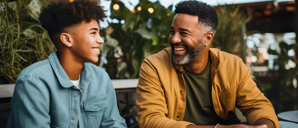 A smiling man and teenager sit together outdoors, facing each other and laughing, surrounded by green plants and warm lights, suggesting a casual, joyful conversation.