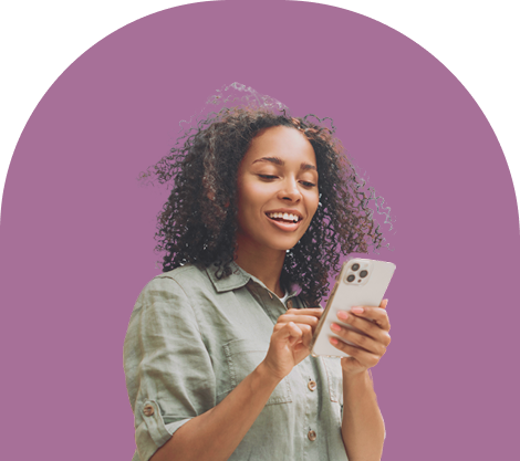 A smiling woman with curly hair wearing a light green button-up shirt looks at and interacts with her smartphone, standing against a solid purple background.